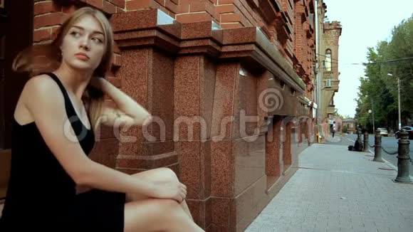迷人的孤独金发女郎穿着黑色的裙子坐在街上视频的预览图