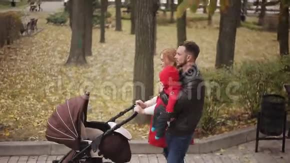 年轻的父母抱着一个孩子带着婴儿车漫步在秋天的公园里视频的预览图