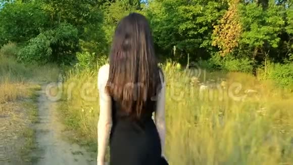 在阳光明媚的日子里一头浓密头发的年轻苗条黑发披着黑色连衣裙在绿色的田野上生长视频的预览图