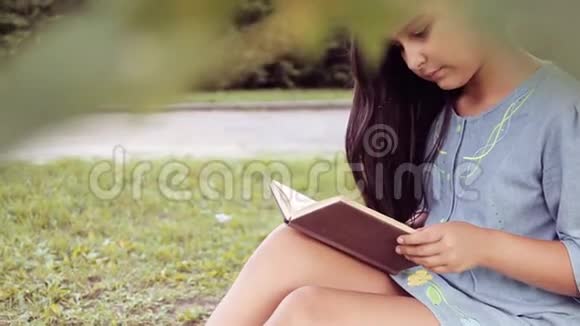 肖像一个留着长发的漂亮小女孩读着一本书坐在树下梦见了一些令人愉快的事情视频的预览图