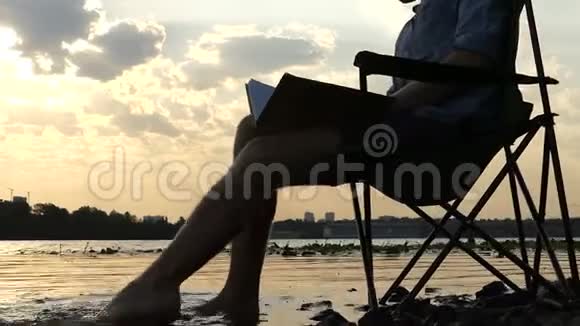 年轻人坐在河岸上写作在水中移动他的脚视频的预览图
