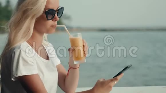 女孩在度假时在酒店咖啡馆喝果汁检查手机以观赏大海和海滩一幅肖像画视频的预览图