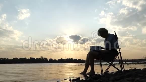 金发碧眼的人坐在河岸上写着把他的脚放在水中视频的预览图