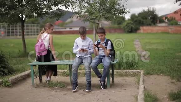 两个男孩和女孩在放学后使用他们的电话可爱的男孩坐在长凳上玩网络游戏视频的预览图