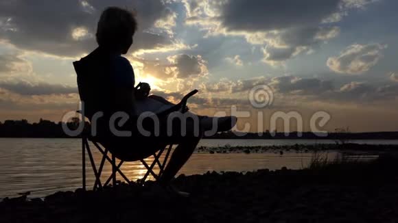 光明的人坐在河岸的椅子上在日落时看书和喝酒视频的预览图