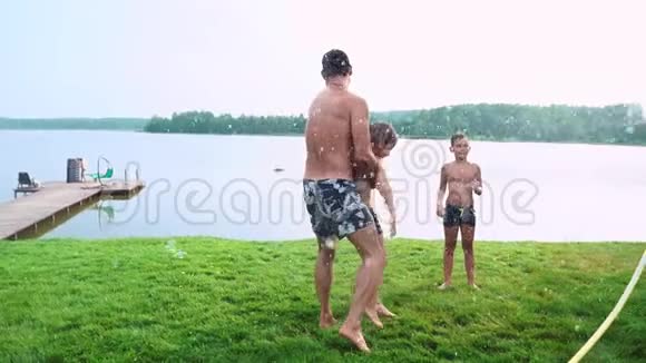 爸爸妈妈和两个儿子在夏天的草地上为草坪浇了一根水管在湖边的乡村别墅视频的预览图