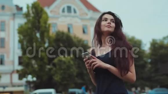 年轻漂亮的黑发女孩站在外面环顾四周等待一个人她用她的手机积极情绪视频的预览图