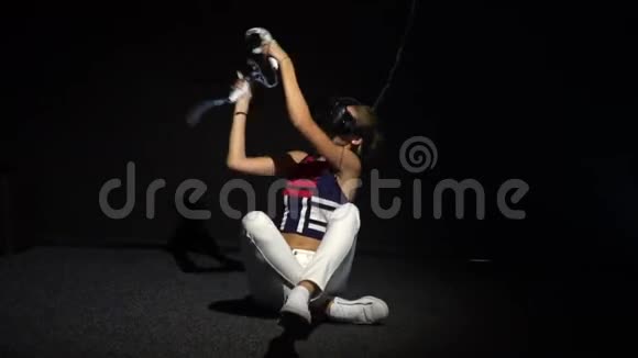 一个身材苗条戴着虚拟现实眼镜的女孩坐在黑暗的房间里的地板上手里拿着操纵杆挥舞着视频的预览图