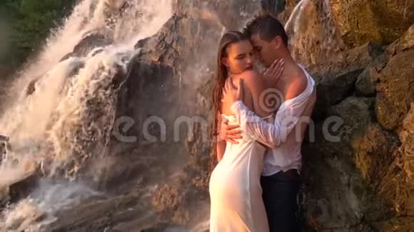日落时分一个男人吸引着一个女孩站在瀑布下的岩石中动作缓慢视频的预览图