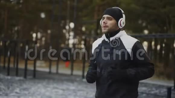 有魅力的男士戴着耳机做热身运动准备在冬季公园听音乐时慢跑视频的预览图