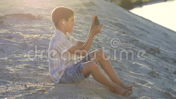 戴着耳机的男孩在智能手机上玩游戏10岁的孩子在日落时分坐在沙滩上享受智能手机视频的预览图