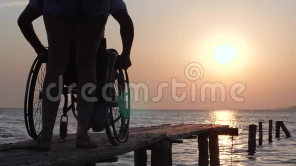 在橙色日落期间协助坐轮椅的残疾人在海上码头行走视频的预览图