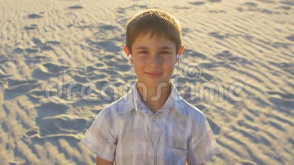 特写男孩在耳机上听音乐十岁的孩子喜欢坐在沙滩背景上的智能手机视频的预览图