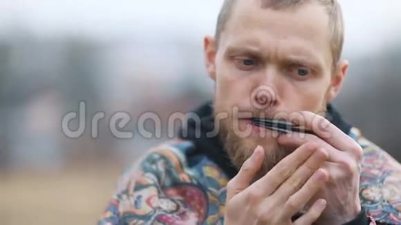 年轻的棕胡子男人在乡村的风景中用唇形乐器演奏富有表现力的视觉视频的预览图