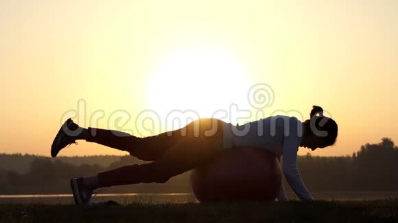 苗条的女人躺在健身球上在日落时抬起一条腿视频的预览图