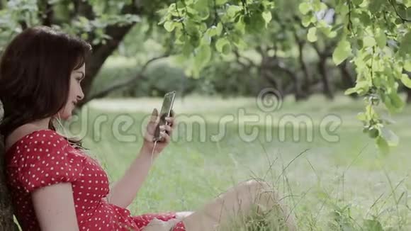 加上坐在树下的草地上的大小女孩使用她的智能手机视频的预览图