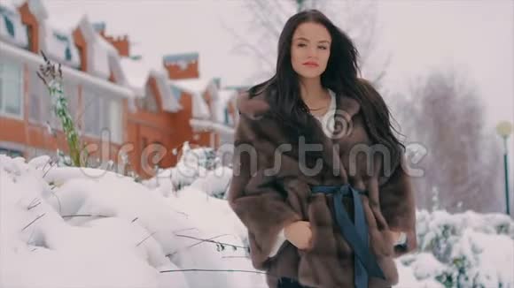 穿着毛皮大衣的布鲁内特女士在冬季大街的建筑物背景上微笑慢动作视频的预览图