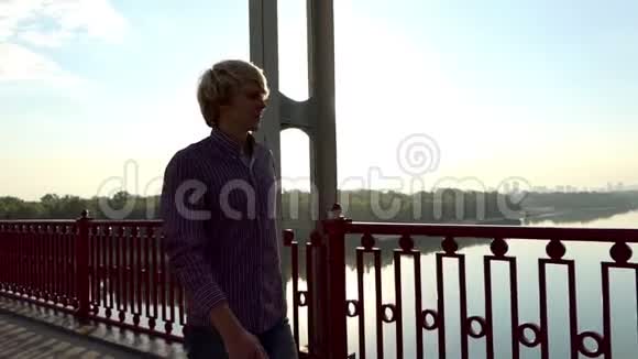 青年男子沿着大桥行进在斯隆莫的日落处拔出他的手机视频的预览图