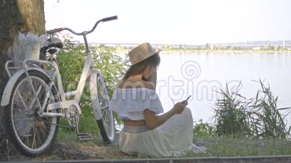 一位美丽的黑发女郎正坐在城市自行车附近的河岸上看一本关于智能手机的书视频的预览图