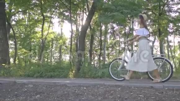 可爱的女孩穿着白色的裙子衬衫在阳光明媚的城市自行车上滚动在公园的小路上享受着阳光明媚的一天视频的预览图