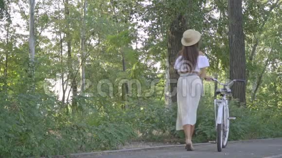 穿着白色裙子的布鲁内特一件衬衫和一顶草帽个阳光明媚的下午公园里手里拿着一辆城市自行车笑着视频的预览图