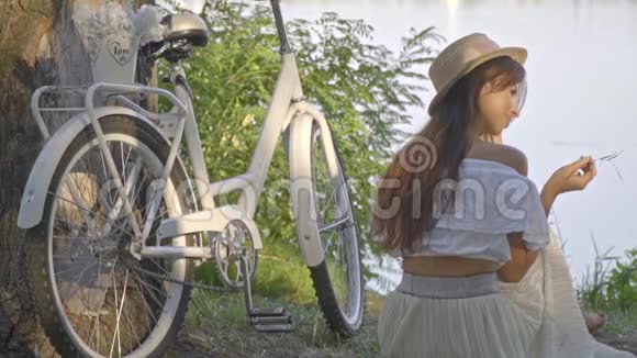 一个浪漫的黑发女人穿着白色长裙衬衫头戴草帽坐在城市自行车旁边的河岸上视频的预览图