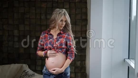 年轻漂亮的孕妇穿着格子衬衫站在靠近窗户的地方慈爱地抚摸着她的腹部未来母亲视频的预览图