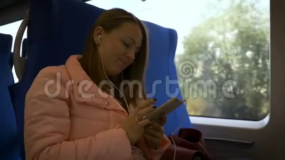 年轻的美女坐在火车上用白色耳机听智能手机里的音乐视频的预览图