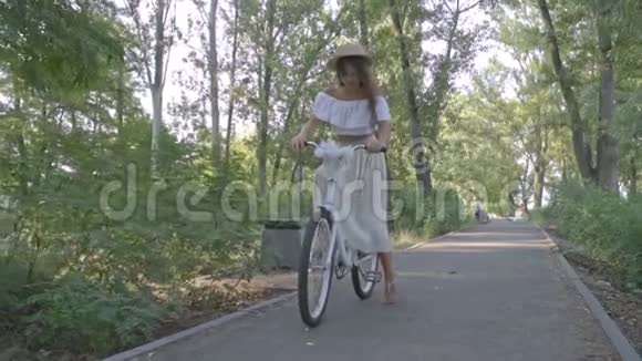 穿着白色裙子上衣和草帽的布鲁内特骑着一辆城市自行车沿着公园的小路慢镜头拍摄视频的预览图