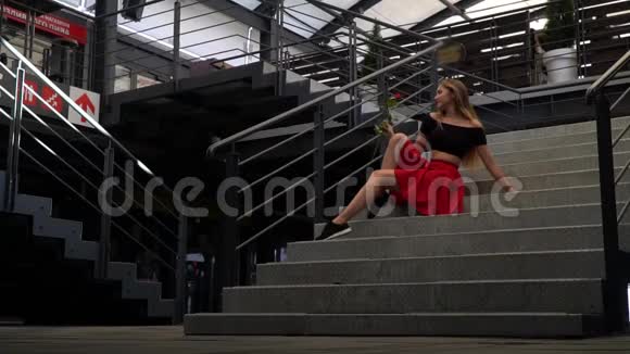 快乐的女孩留着长发坐在商场的楼梯上欣赏着手中的红玫瑰视频的预览图