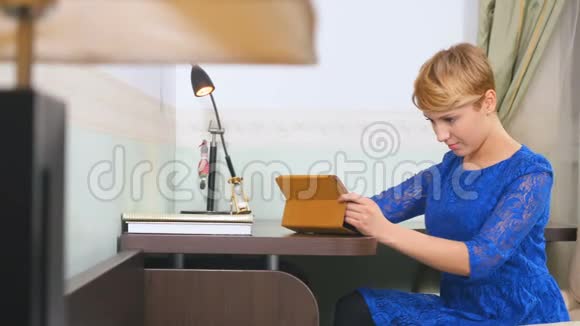穿蓝衣服的年轻女子在家里用平板电脑坐在桌旁视频的预览图