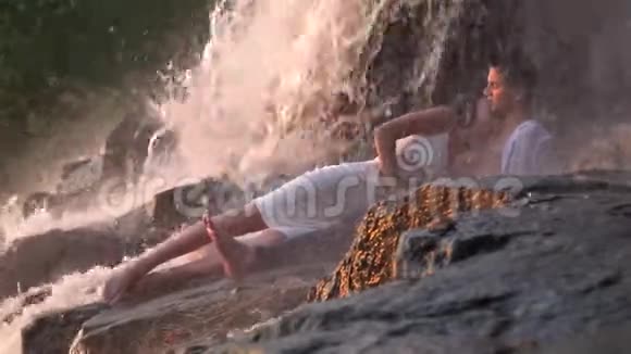 一个穿着裙子的女孩亲吻一个男人的脖子当他们躺在瀑布下的岩石上时他们会掉下来视频的预览图