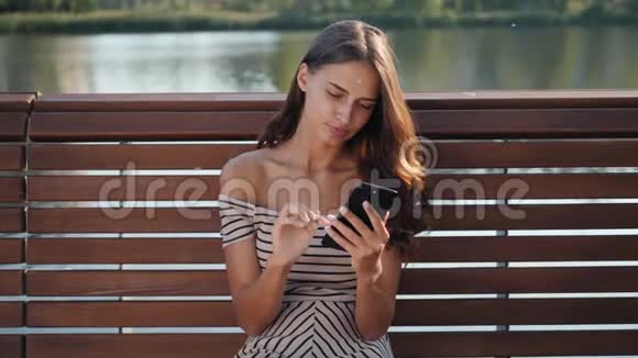 快乐女学生坐在城市公园的长凳上用智能手机年轻女子微笑着用应用程序长时间视频的预览图
