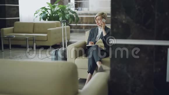 金发碧眼的女商务人士坐在酒店大堂的扶手椅上用手机说话写着记事本微笑着视频的预览图