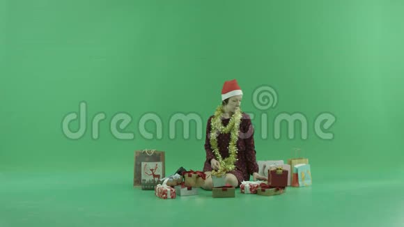 一位年轻女子正坐在绿色的屏幕上在自己身边打开圣诞礼物视频的预览图