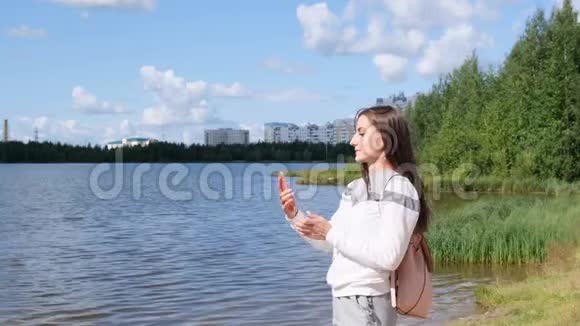 带背包的漂亮女孩在湖边自拍并在Skype上用手机交谈视频的预览图