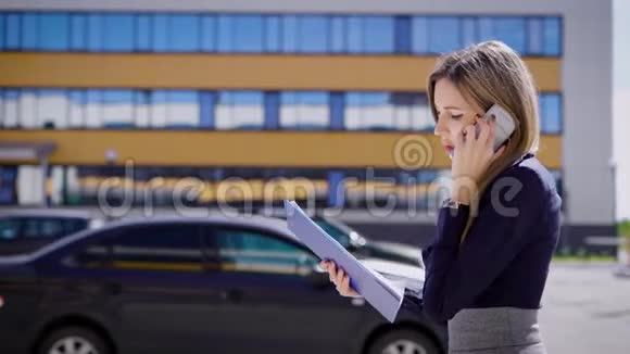 一位忙碌的商务女士拿着重要的合同在手机上交谈她走在停车场旁那里有汽车视频的预览图