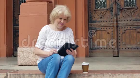 老妇人坐在台阶上用平板电脑和喝咖啡背景中的老式建筑视频的预览图