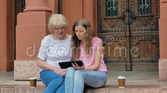 年长的女人和年轻的女孩坐在台阶上使用智能手机和喝咖啡背景中的老式建筑视频的预览图