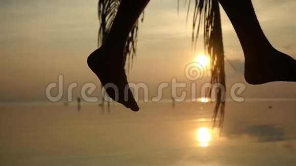 把挥舞着的男腿合上在令人惊叹的日落中年轻人坐在热带海滩的棕榈树上慢动作视频的预览图