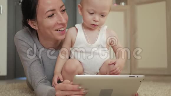 年轻的母亲和小男孩在地板上的地毯上玩平板电脑妈妈和儿子玩数字平板电脑视频的预览图