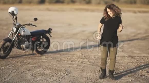 年轻漂亮的女人在日落或日出时在沙漠里跳舞然后坐在摩托车上骑着车离开慢动作视频的预览图