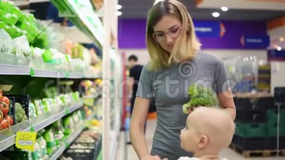 年轻的母亲正在选择卷心菜和其他蔬菜而她的孩子坐在购物车里家庭视频的预览图