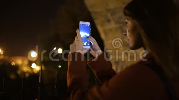 迷人的年轻女性在夜间漫步街头时在智能手机上拍照慢动作视频的预览图