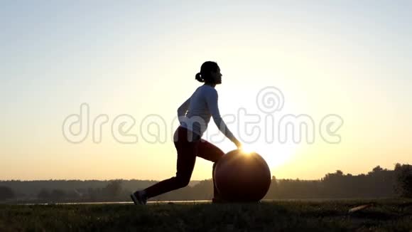 苗条的女人站着跪着在邋遢的夕阳下滚动她的健身球视频的预览图