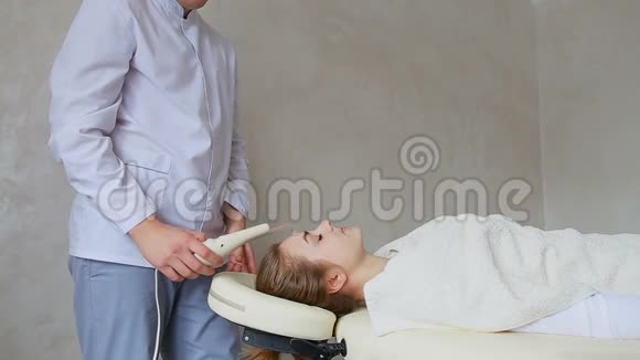 男专家化妆师在躺在沙发上的病人女孩的背景下演示面部按摩装置的工作视频的预览图