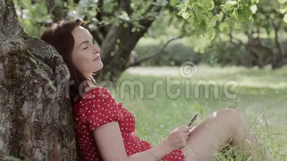 加上坐在树下的草地上的大小女孩使用她的智能手机视频的预览图