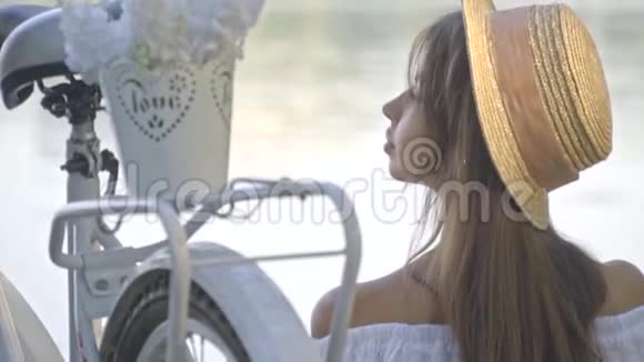 一位深谋远虑的黑发女子戴着草帽穿着白色衬衫坐在她的城市自行车旁边欣赏着这条河视频的预览图