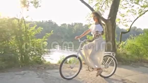 一个穿着长裙一件白色衬衫挥舞着草帽的可爱黑发女子骑着白色的城市自行车在河边的路上骑着视频的预览图