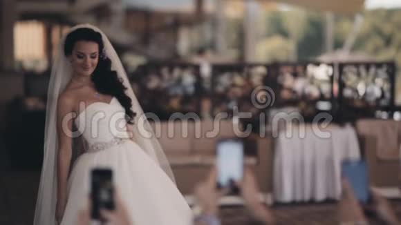美丽的新娘在她的朋友在智能手机上拍照时摆姿势结婚那天穿白色裙子的幸福女人视频的预览图